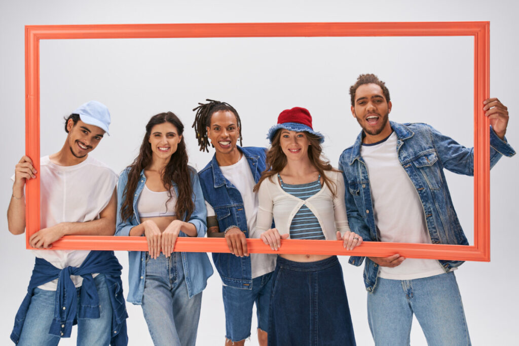 Group portrait of stylish interracial friends posing with picture frame on grey backdrop, fashion